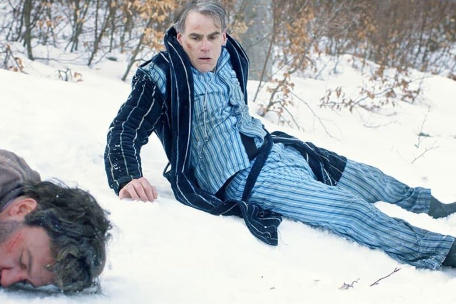 Der Protagonist sitzt nach einem Sturz mit überraschtem Gesichtsausdruck im Schnee, nur in einem Pyjama gekleidet. Neben ihm liegt ein scheinbar bewusstloser Mann.
