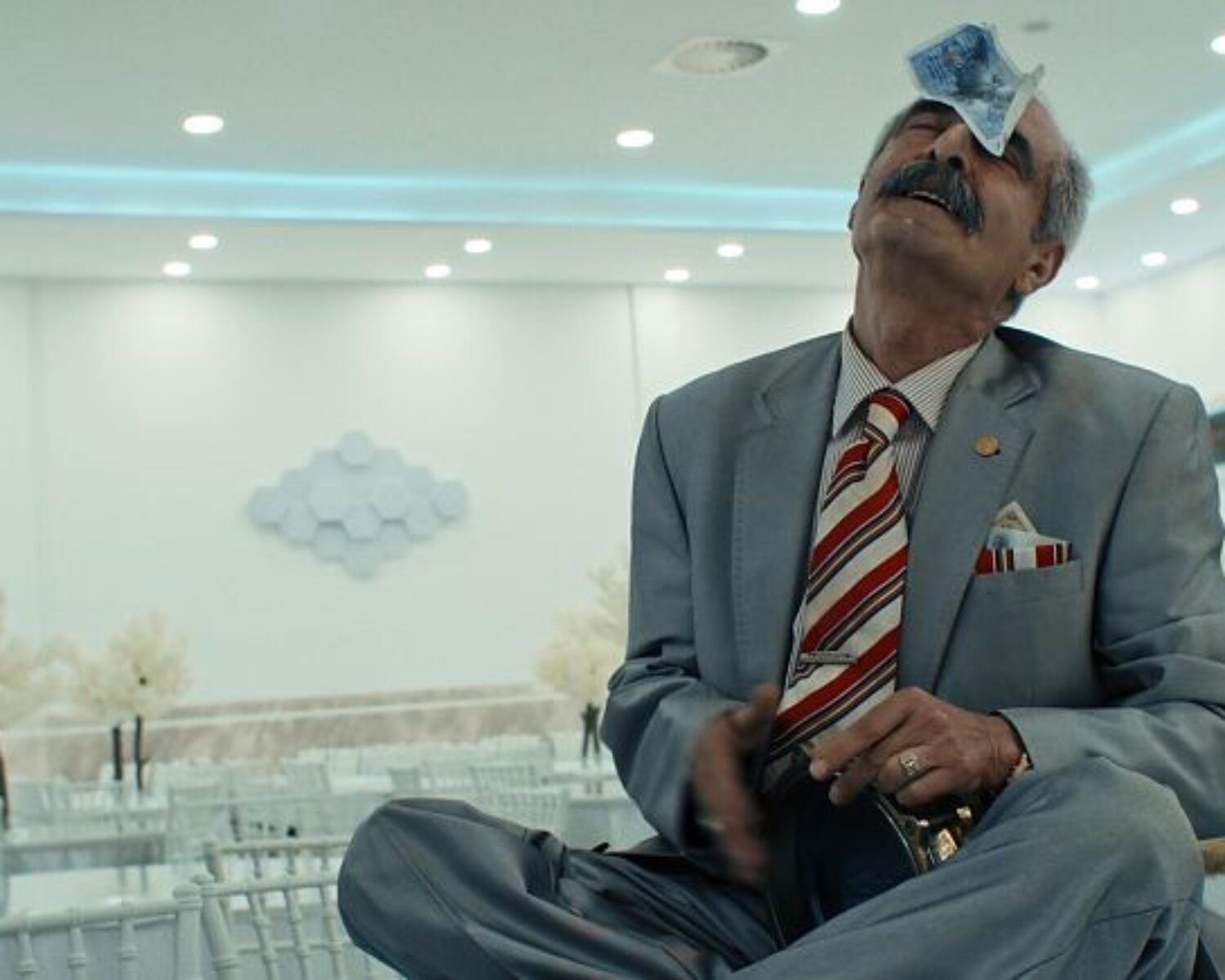 Ein fein gekleideter, älterer Mann sitzt auf einem Tisch und hat einen Geldschein auf die Stirn geklebt. Es handelt sich um eine Szene aus dem Film.