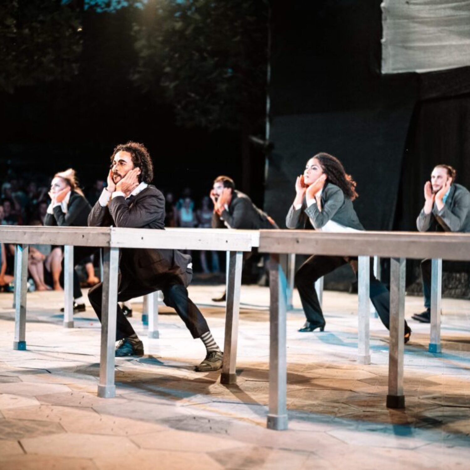 Fünf Schauspielende während der Performance, die Arme auf jeweils einen Schreibtisch gelehnt. Im Hintergrund sitzt gespanntes Publikum.